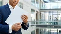 Middle-aged male professional in dark business suit holding blank white document in modern glass office building interior with natural light.