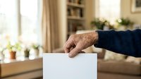 Senior male voter with aged hands casting ballot in white box during election, home interior with warm lighting and bookshelf background.