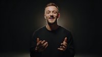 Young Caucasian man in black shirt gesturing expressively while speaking against dark background, conveying enthusiasm and engagement during presentation or interview.