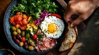Vibrant breakfast bowl with sunny side up egg, fresh vegetables, prosciutto, olives and sourdough bread on rustic wooden table. Fresh morning meal concept.