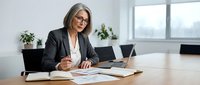 Mature woman with gray hair and glasses wearing black blazer working at desk with laptop and documents in modern office environment.