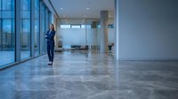 Professional woman in business attire standing in modern empty office lobby with polished marble floors, floor-to-ceiling windows, and contemporary architecture for corporate workspace concepts.