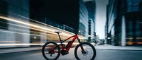 Red electric mountain bike stationary on urban street with motion-blurred skyscrapers and traffic, showcasing modern e-bike technology for city commuting and sustainable transportation.
