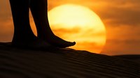 Silhouetted feet standing on wooden surface with large golden sun setting on horizon, creating warm orange sky backdrop for travel, nature, and lifestyle concepts.
