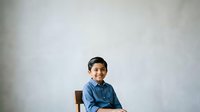 Young boy with dark hair and South Asian ethnicity, wearing a blue denim shirt, sitting on a wooden chair against a neutral gray background. Portrait suitable for education, family, and lifestyle content.