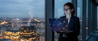 Woman in business attire holding laptop with digital network visualization, standing by high-rise window overlooking illuminated cityscape at dusk for corporate technology concepts.