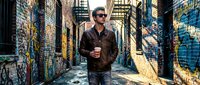 Young Caucasian man in leather jacket and sunglasses walking through urban alley with colorful graffiti street art, holding coffee cup in city environment.