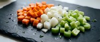 Fresh diced vegetables including orange carrots, white onions and green celery arranged on dark slate board for cooking preparation and meal planning.