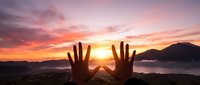 Hands reaching toward vibrant sunset over mountain landscape with dramatic orange and pink sky clouds creating inspirational freedom concept.