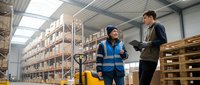 Two warehouse workers discussing logistics operations in modern distribution center with pallet racks and industrial equipment for supply chain management.