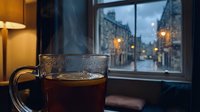 Steaming hot coffee mug on windowsill overlooking cozy evening city street with warm glowing lights creating peaceful indoor atmosphere for relaxation and comfort.