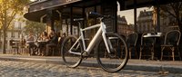 White fixed-gear bicycle parked on cobblestone street outside urban cafe with people dining, golden hour lighting, European city setting for lifestyle and travel content.