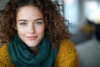 Young woman with natural curly brown hair smiling warmly at camera, wearing cozy yellow knit sweater and green scarf. Genuine happiness and confidence in casual winter fashion.
