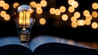 Glowing vintage light bulb on open book against blurred golden bokeh background creates atmospheric mood for reading and inspiration.