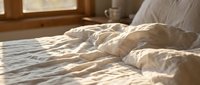 Unmade bed with rumpled beige bedding and pillows in warm morning sunlight, suitable for lifestyle, home decor, and bedroom design projects.