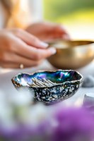Iridescent abalone shell with vibrant blue and purple mother of pearl interior on white surface, sage burning ritual setup with blurred background and soft natural lighting.