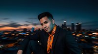 Young Hispanic businessman in orange shirt and dark jacket posing confidently against blurred city skyline at dusk with warm lighting for corporate marketing.