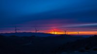 Wind turbines silhouetted against dramatic sunset sky with orange and purple clouds over mountain landscape for renewable energy concepts.