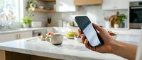 Hand holding smartphone with blank screen in modern kitchen interior with white countertops and natural lighting for app mockup and digital lifestyle concepts.