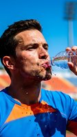 Adult male athlete with dark hair and facial hair, Middle Eastern or Mediterranean ethnicity, drinking water from bottle during outdoor sports training under bright sunlight.