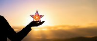 Silhouetted hand holding glowing star against golden sunset sky with mountains, representing achievement, success, hope and aspirations for business and personal growth.