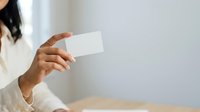 Woman with dark hair and medium skin tone holding blank white business card in her hand, wearing light beige sweater, isolated on white background for corporate branding and professional design use.