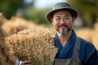 Mature Asian farmer with gray beard wearing straw hat and overalls holds freshly harvested wheat bundle while smiling at camera in golden wheat field during sunset.