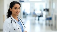 Professional Hispanic female doctor in white coat with stethoscope smiling confidently in modern hospital corridor for healthcare marketing materials.