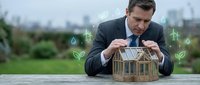 Businessman in dark suit and glasses examining wooden house model with green energy icons and sustainability symbols overlaid, urban skyline background, representing eco-friendly real estate development.