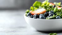 Fresh mixed salad with ripe blueberries, strawberries and green lettuce leaves in white ceramic bowl on light marble background with copy space.