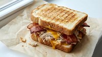 Grilled sandwich with crispy bacon, melted cheese and caramelized onions on toasted bread, served on parchment paper for breakfast or lunch meal.
