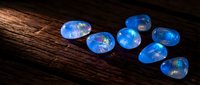 Blue opal gemstones with iridescent fire scattered on dark wooden surface for jewelry making and luxury accessories design projects.