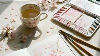 Watercolor painting workspace with cherry blossom tea cup, pink palette, brushes and scattered petals on white surface for artistic inspiration.