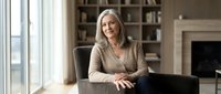 Confident mature businesswoman with gray hair sitting at desk in modern office interior with bookshelf and fireplace, professional corporate executive portrait.