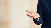 Caucasian male professional in dark business suit gesturing with open palm hand in welcoming or presenting pose against neutral beige background for corporate communication.