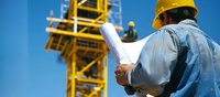 Male construction supervisor in hard hat reviewing blueprints at building site with yellow crane and worker in background against blue sky. Industrial development scene.
