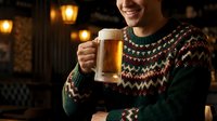 Man in festive sweater holding beer mug indoors at night, celebrating holidays with alcoholic beverage in warm cozy setting.