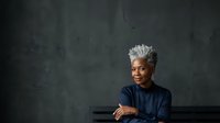 Mature woman with short white hair and dark skin wearing navy blue shirt leaning on table against textured gray wall background, professional portrait for business and corporate use.