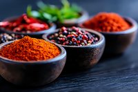 Vibrant red chili powder and mixed peppercorns in rustic wooden bowls against dark background, selective focus on aromatic spices for cooking and seasoning.