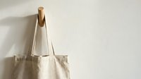 Natural linen tote bag hanging on wooden peg against neutral wall background for sustainable shopping and eco-friendly lifestyle concepts.
