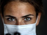 Close up portrait of young woman with freckles and intense eyes wearing protective medical face mask against dark background, healthcare concept.
