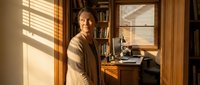 Senior African American woman working at wooden desk in warm home office with natural sunlight streaming through window blinds creating shadow patterns.