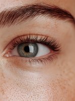 Close-up macro shot of gray-green eye with long natural eyelashes and light freckles on fair skin, showing fine details of iris pattern and texture.