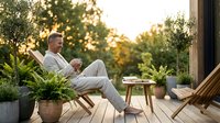 Mature man relaxing on wooden deck with coffee during golden hour sunset, enjoying peaceful outdoor lifestyle moment surrounded by lush green plants and garden.