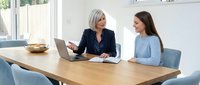 Professional mature businesswoman mentoring young female colleague during office meeting with laptop and documents on wooden conference table.