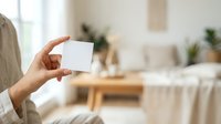 Person holding blank white card in modern bedroom interior with natural light and minimalist decor, suitable for business card mockups and promotional designs.