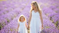 Young blonde mother and daughter holding hands walking through purple lavender field during golden hour sunset wearing matching white dresses for family bonding.