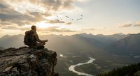Male hiker with backpack sitting on rocky cliff edge reading map during golden hour sunrise over mountain valley with river and flying birds.