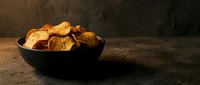 Crispy golden potato chips in dark bowl on rustic wooden surface with dramatic lighting for snack food advertising and culinary presentations.