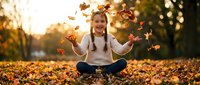 Young girl with braids sitting cross-legged throwing colorful autumn leaves in the air during golden hour sunset in park with warm bokeh background.